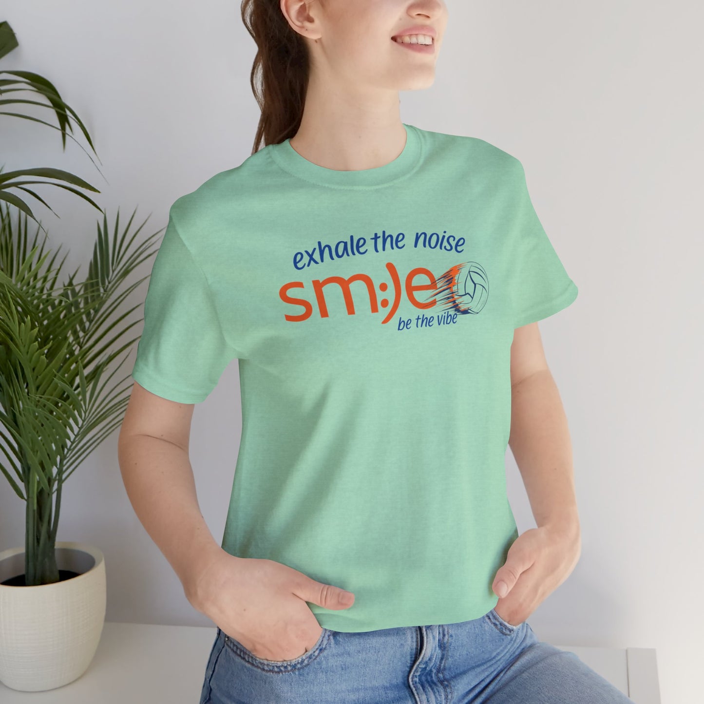 Person wearing a light green t-shirt with motivational text, standing indoors next to a plant.